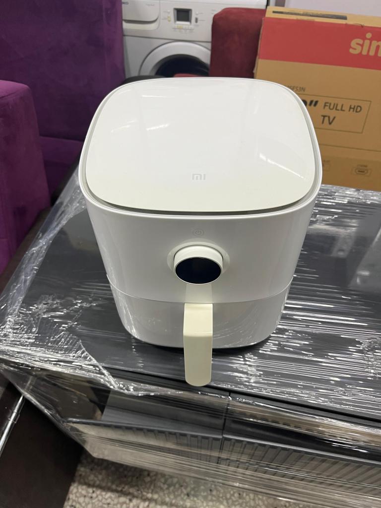 Bir beyaz hava fritözü, masanın üstünde paketlenmiş şekilde duruyor. Arkada çeşitli ev aletleri ve televizyon kutusu görünmekte.